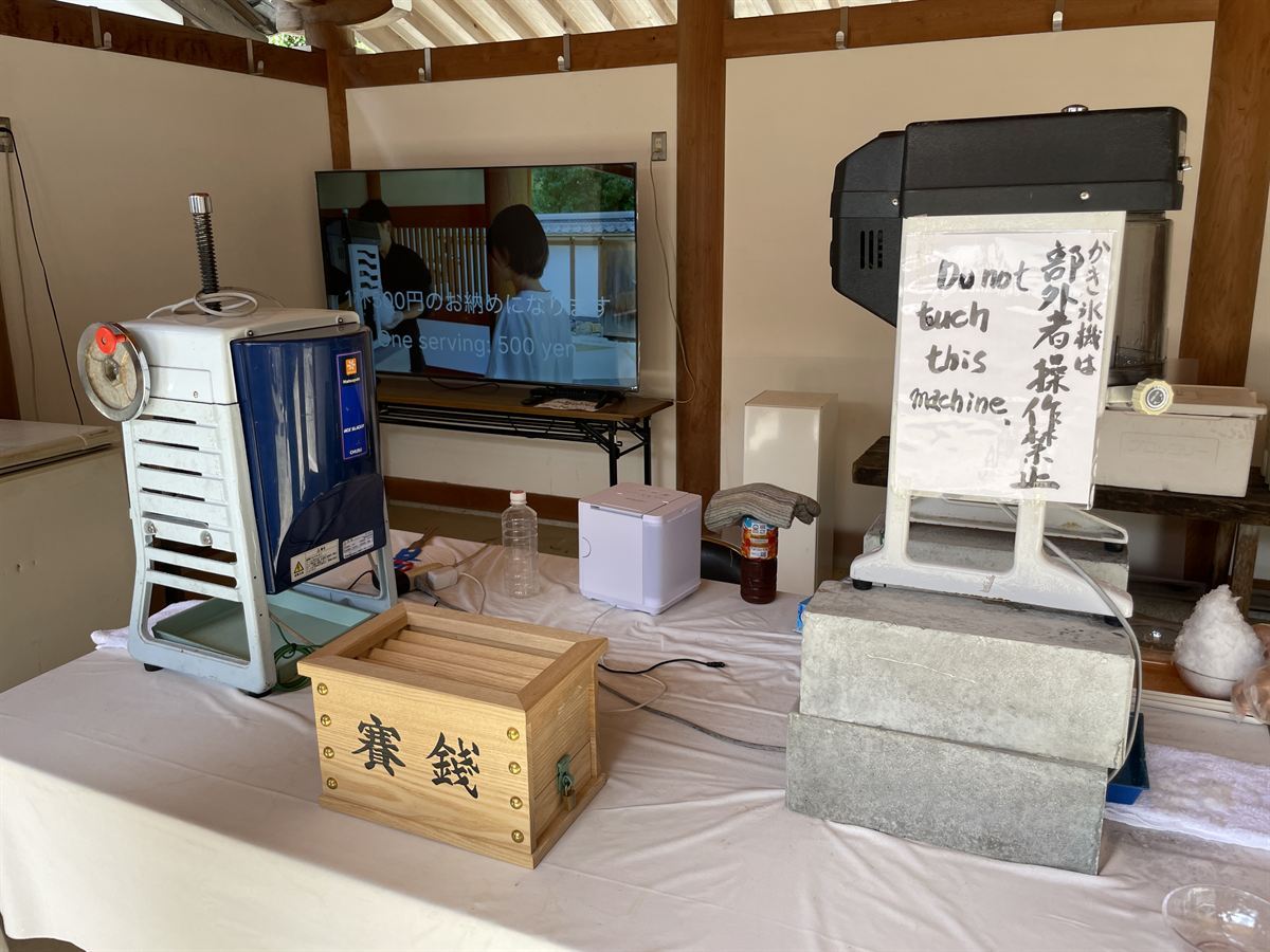 かき氷献氷の受付。業務用のかき氷機が2台、木の賽銭箱、作法の動画を流すモニターが並ぶ