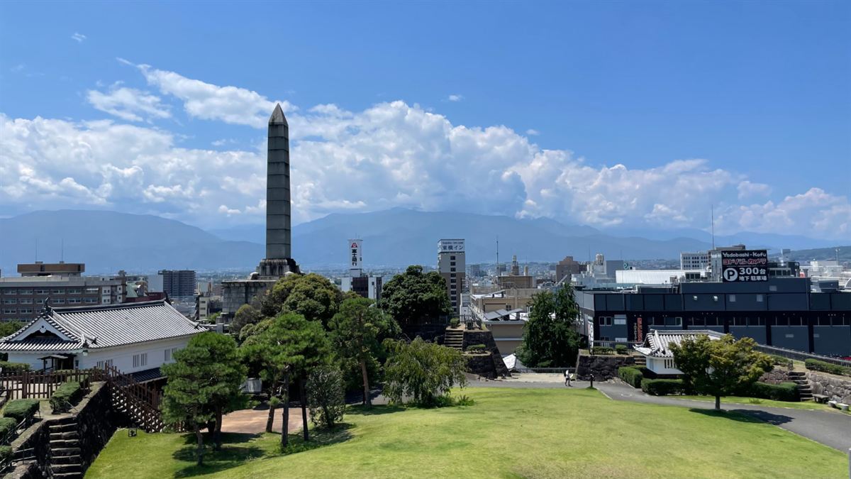 甲府城跡の園内。芝生の広場と松、白壁の建物、奥に大きな石塔と甲府の市街地、その向こうに山並み