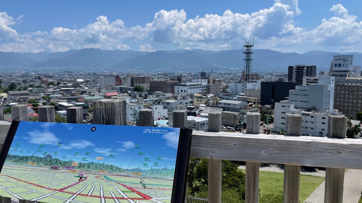 甲府城跡の天守台から南を見た眺め。市街地の向こうに山並みが連なり、手前に眺望の案内板が置かれている