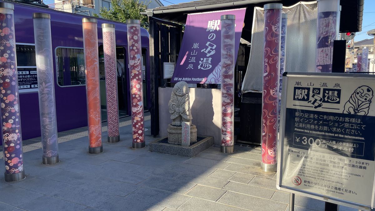 嵐電嵐山駅の「駅の足湯」の入口と料金看板。奥に紫色の嵐電の車両とキモノフォレストの柱