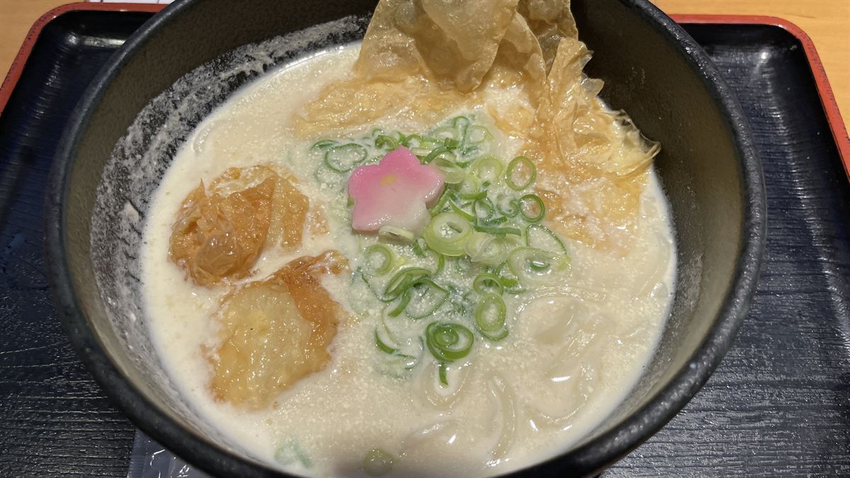 黒い丼に入ったゆばチーズうどん。湯葉とねぎ、花形のかまぼこがのっている