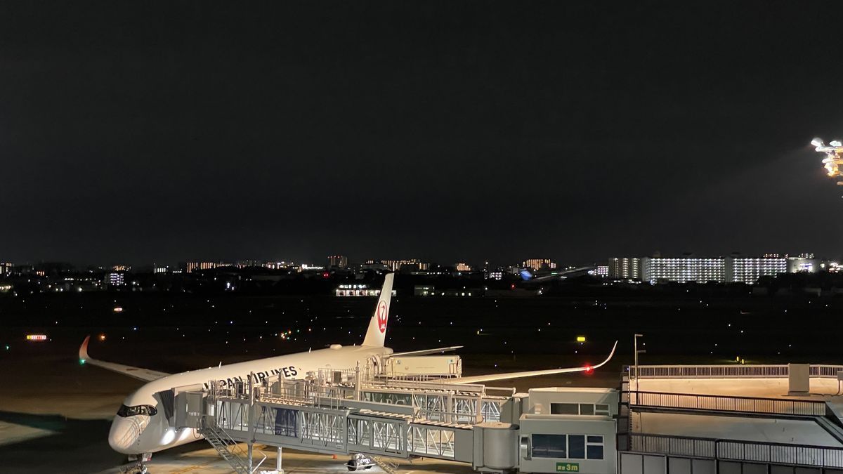 夜の展望デッキから見た、搭乗口に止まっているJAPAN AIRLINESの飛行機。ボーディングブリッジと照明、遠くに街の灯りが見える