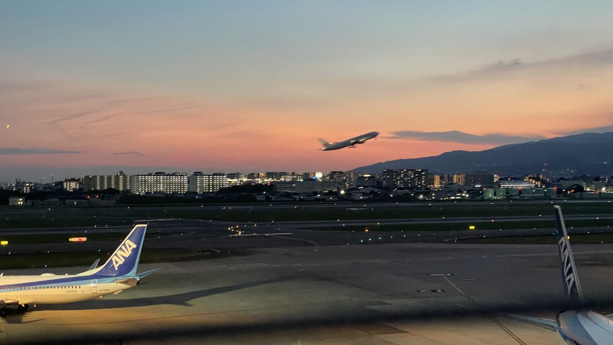夕焼けの空へ離陸していく飛行機。手前にANAの飛行機の尾翼、奥に山並みと街の灯りが見える