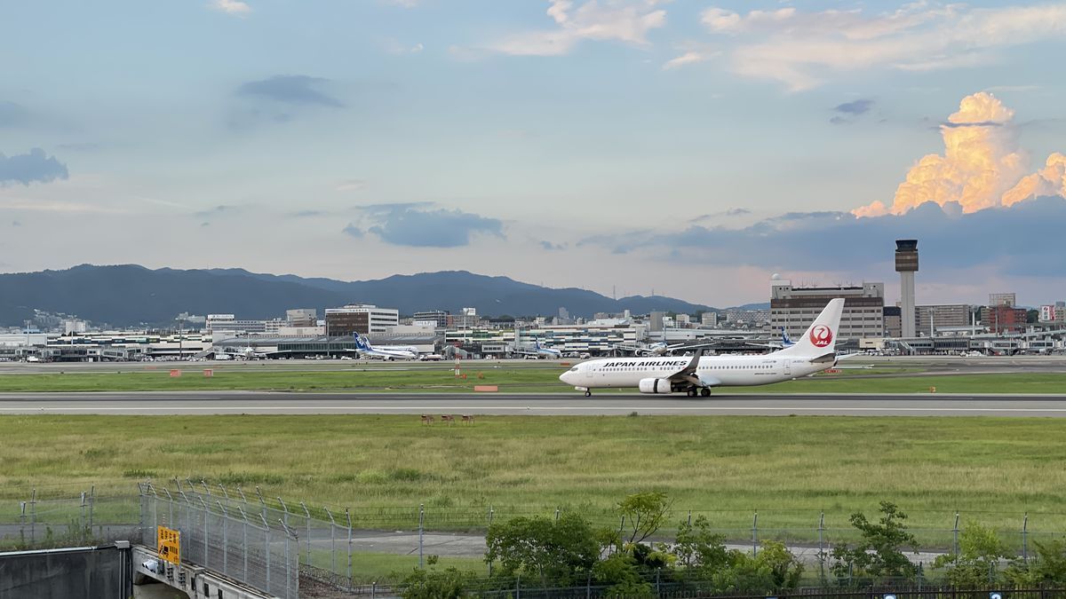 夕方の滑走路を走るJAPAN AIRLINESの飛行機。奥に空港のターミナルと管制塔、山並みと、夕日に照らされた雲が見える