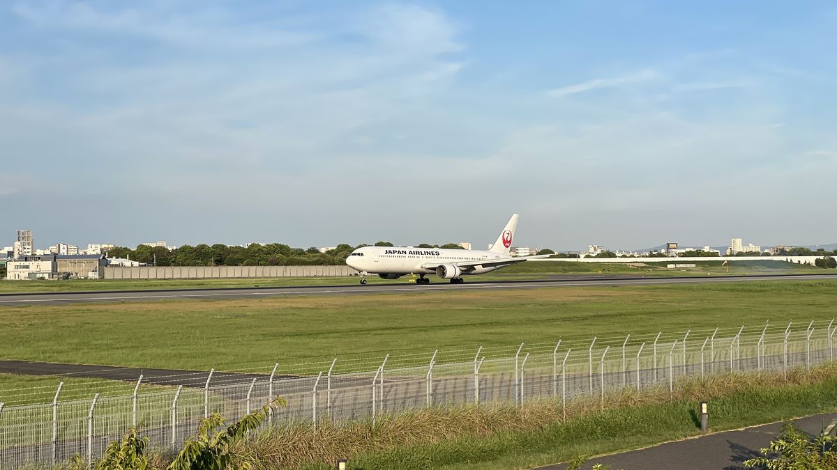 滑走路を走るJAPAN AIRLINESの大きな機体。手前に空港のフェンス、奥に街並みと空が見える