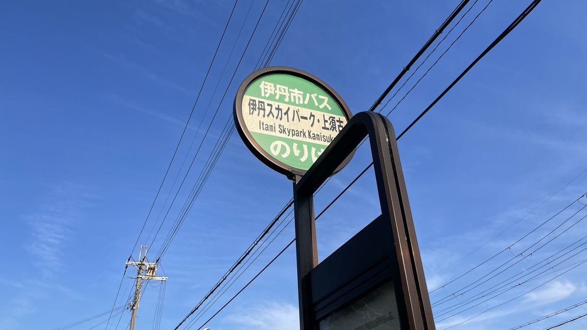 伊丹市バスのバス停の標識。「伊丹スカイパーク・上須古」と書かれ、うしろに青空と電線が見える