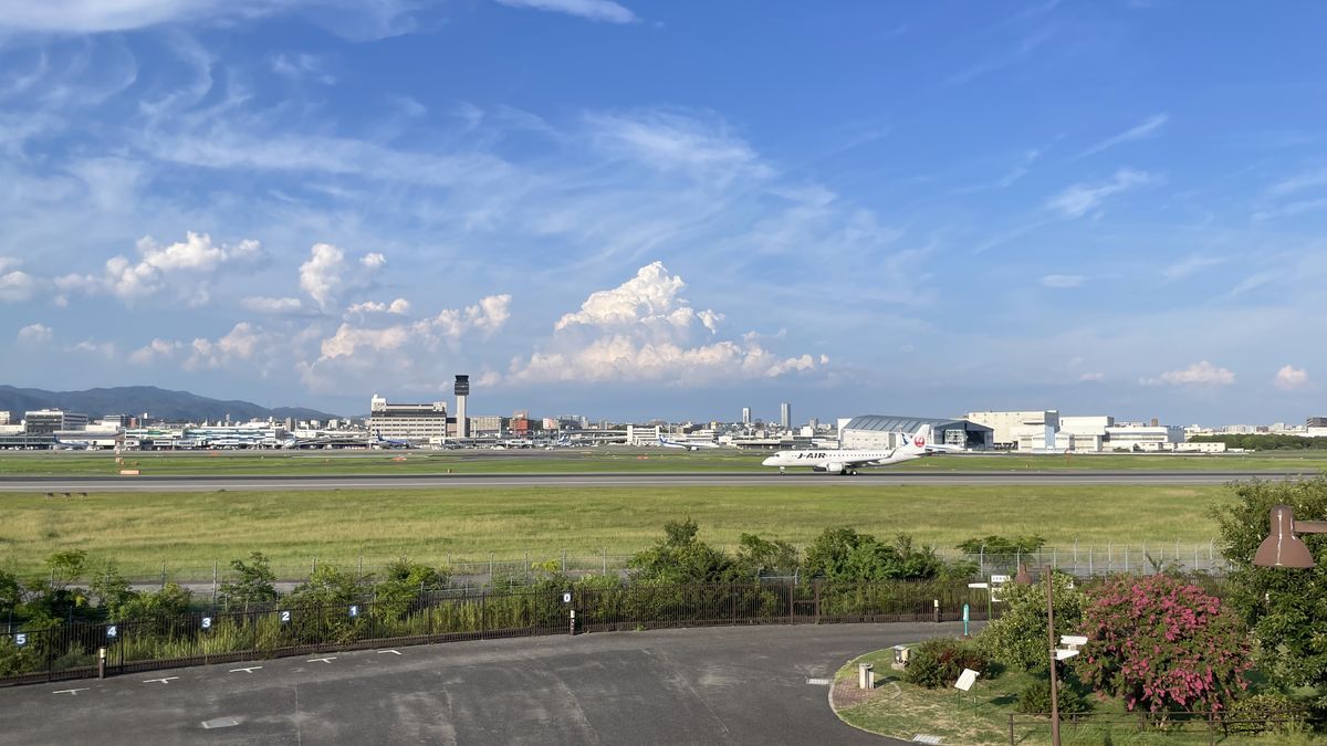 伊丹スカイパークから見た、滑走路を走るJ-AIRの飛行機。奥に空港のターミナルと管制塔、手前に駐車場と植え込み、ピンクの花の木がある