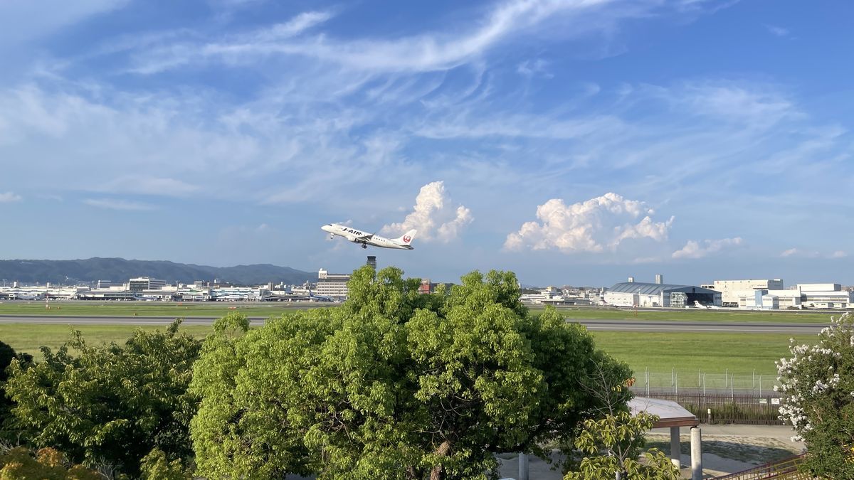 離陸して空へ上がっていくJ-AIRの飛行機。手前に木々、奥に滑走路と空港の建物、青空と白い雲が見える