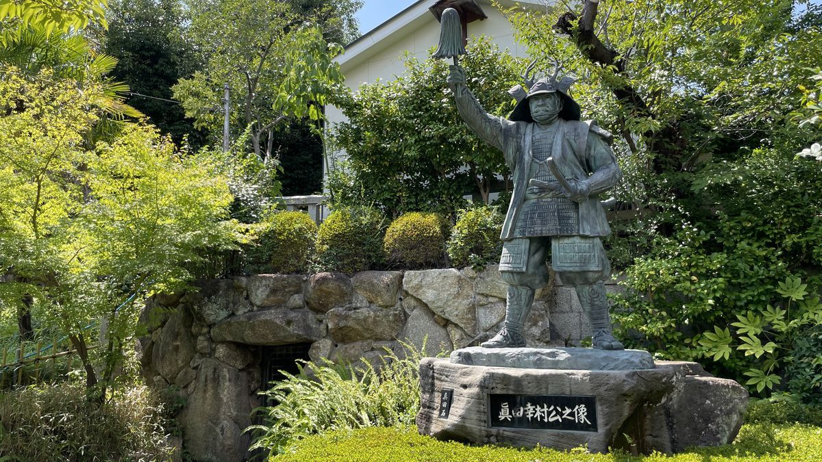 三光神社の真田幸村公之像。甲冑姿で右手を高く上げた銅像の台座に「真田幸村公之像」とあり、左の石組みの奥に穴の入口が見える
