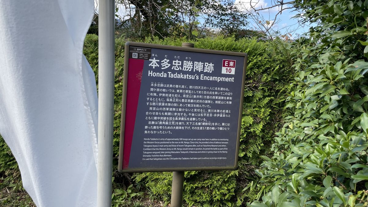 本多忠勝陣跡の案内板「本多忠勝陣跡 Honda Tadakatsu's Encampment」。うしろに生け垣、左に白いのぼりが見える