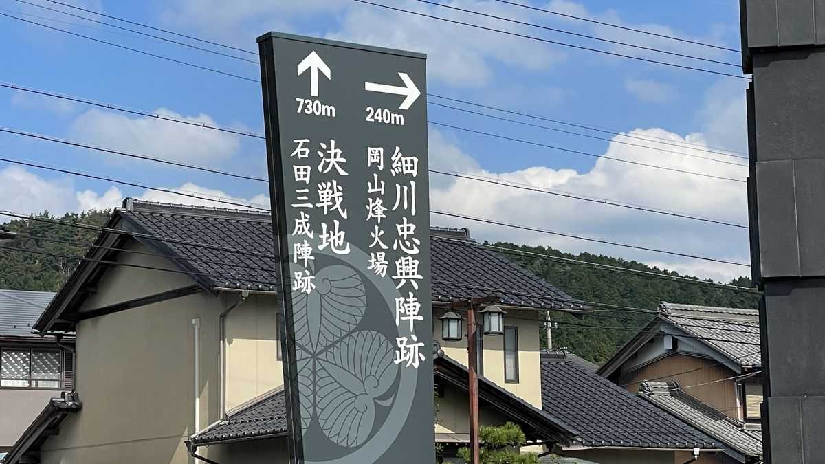 道ばたの道しるべ。「決戦地 石田三成陣跡 730m」「細川忠興陣跡 岡山烽火場 240m」と矢印で書かれ、うしろに家の屋根と山が見える