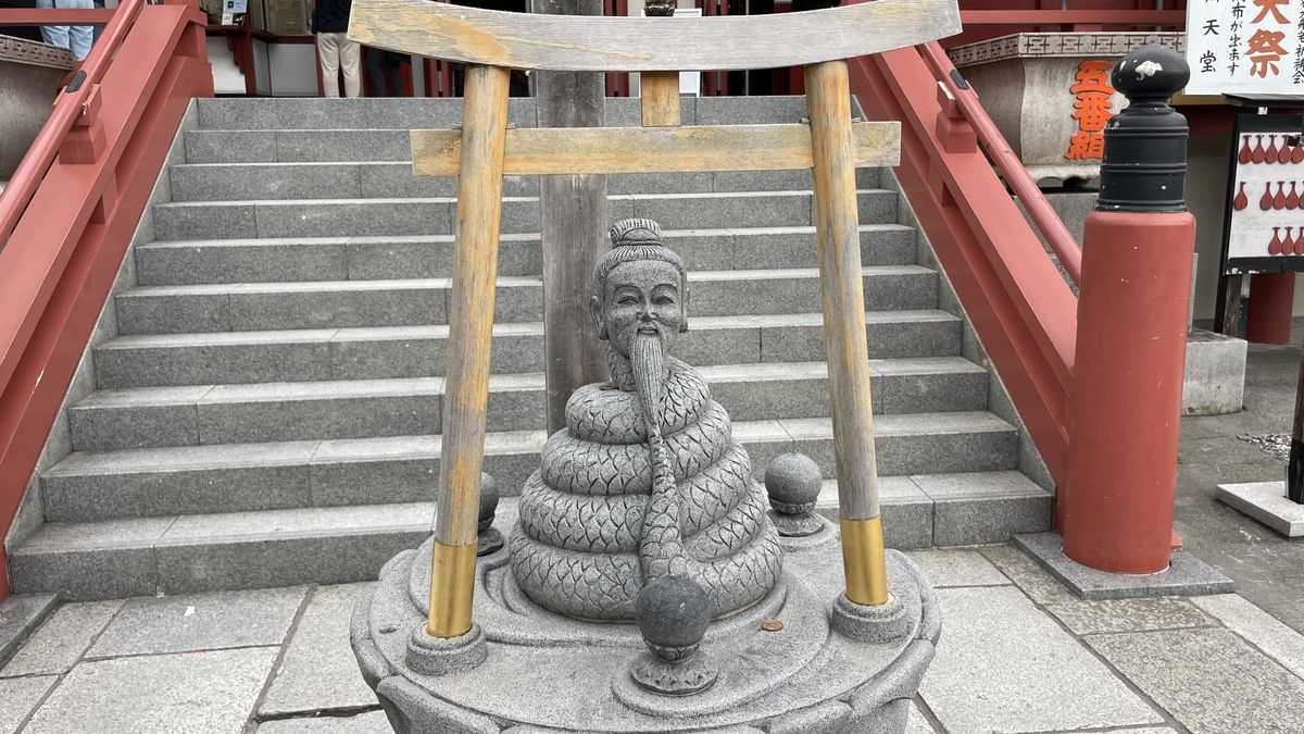 不忍池辯天堂の石段の前の石像。小さな鳥居の下に、とぐろを巻いた蛇の体と、長いひげの老人の顔がある