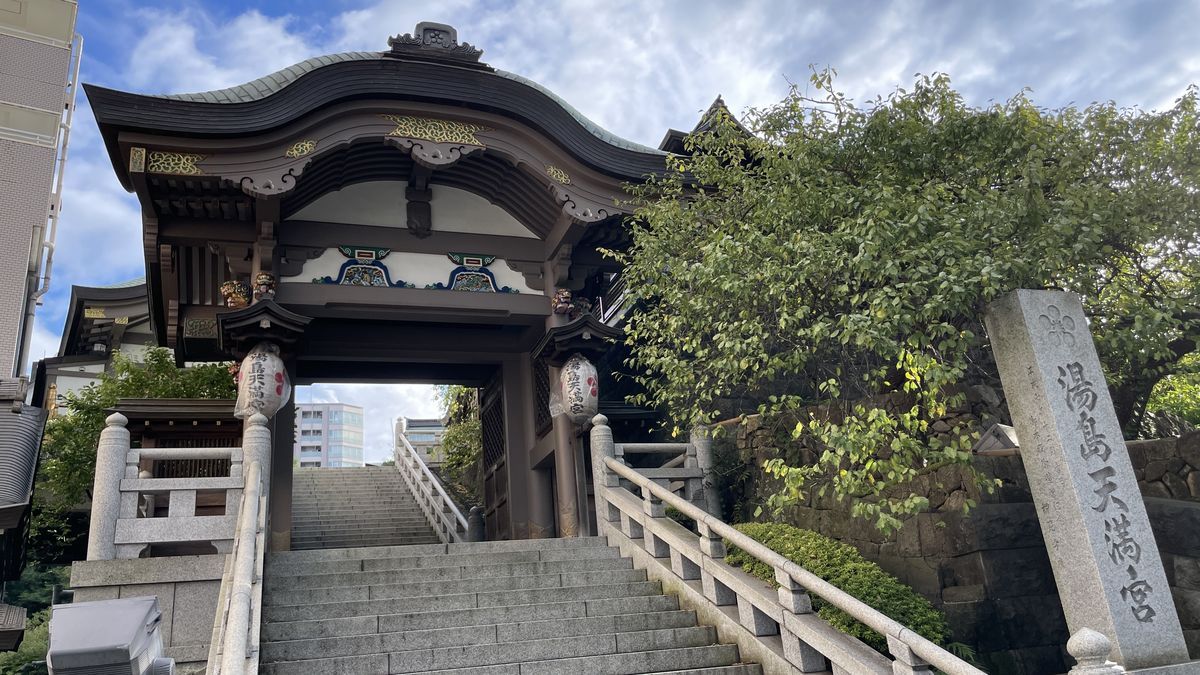 湯島天満宮の門と石段。右に「湯島天満宮」と刻まれた石柱が立ち、空は晴れている