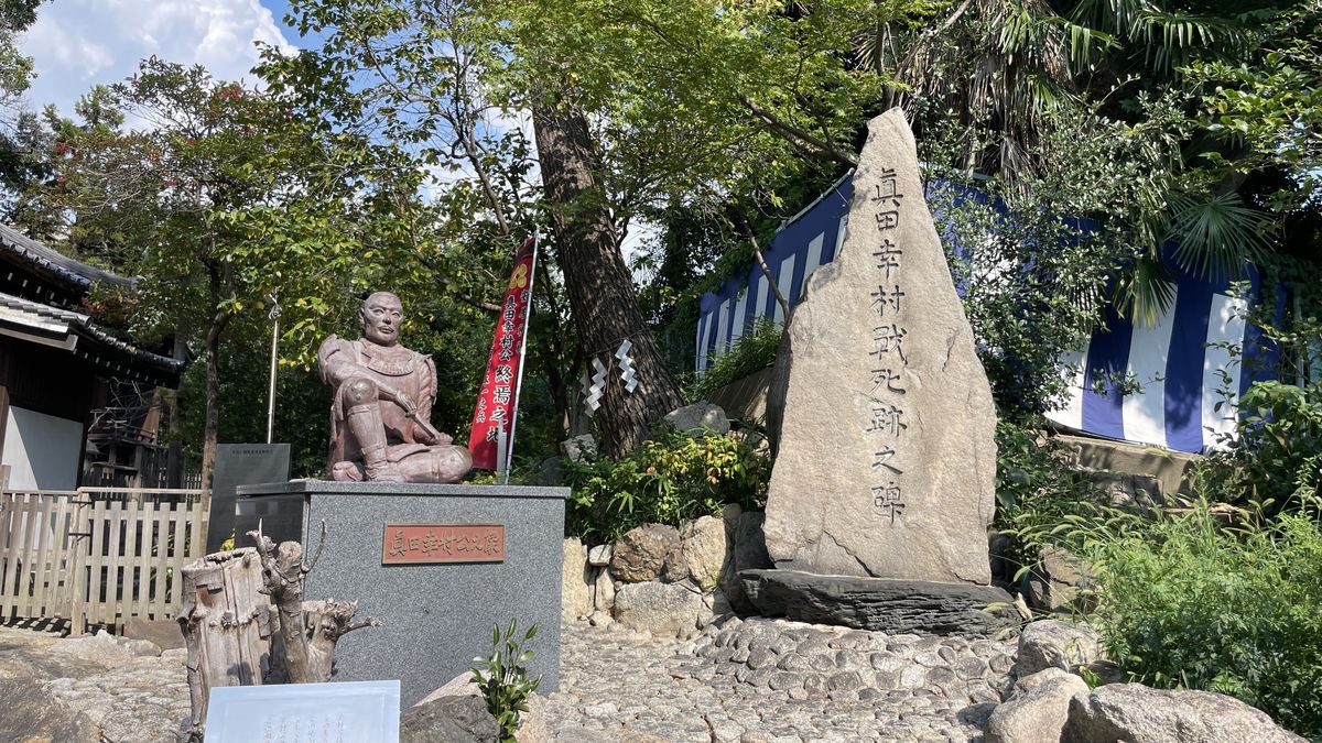安居神社の真田幸村公の座像と「真田幸村戦死跡之碑」。右の石碑に碑文が刻まれ、まわりに木々が茂っている